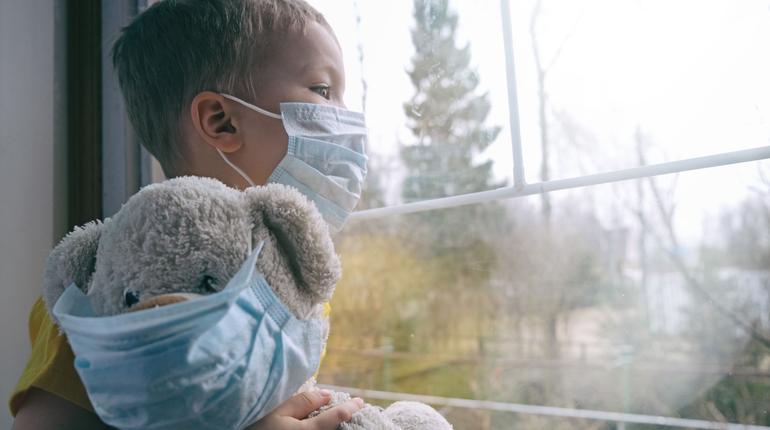 Sad illness child on home quarantine. Boy and his teddy bear both in protective medical masks sits on windowsill and looks out window. Virus protection, coronavirus pandemic, prevention epidemic.